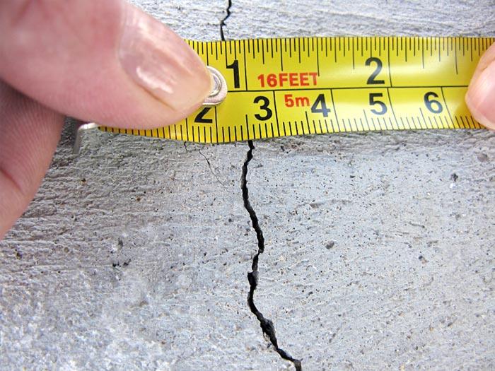 Hand Holding A Measuring Tape Against A Cracked Concrete Basement Foundation Wall, Illustrating The Size Of The Crack