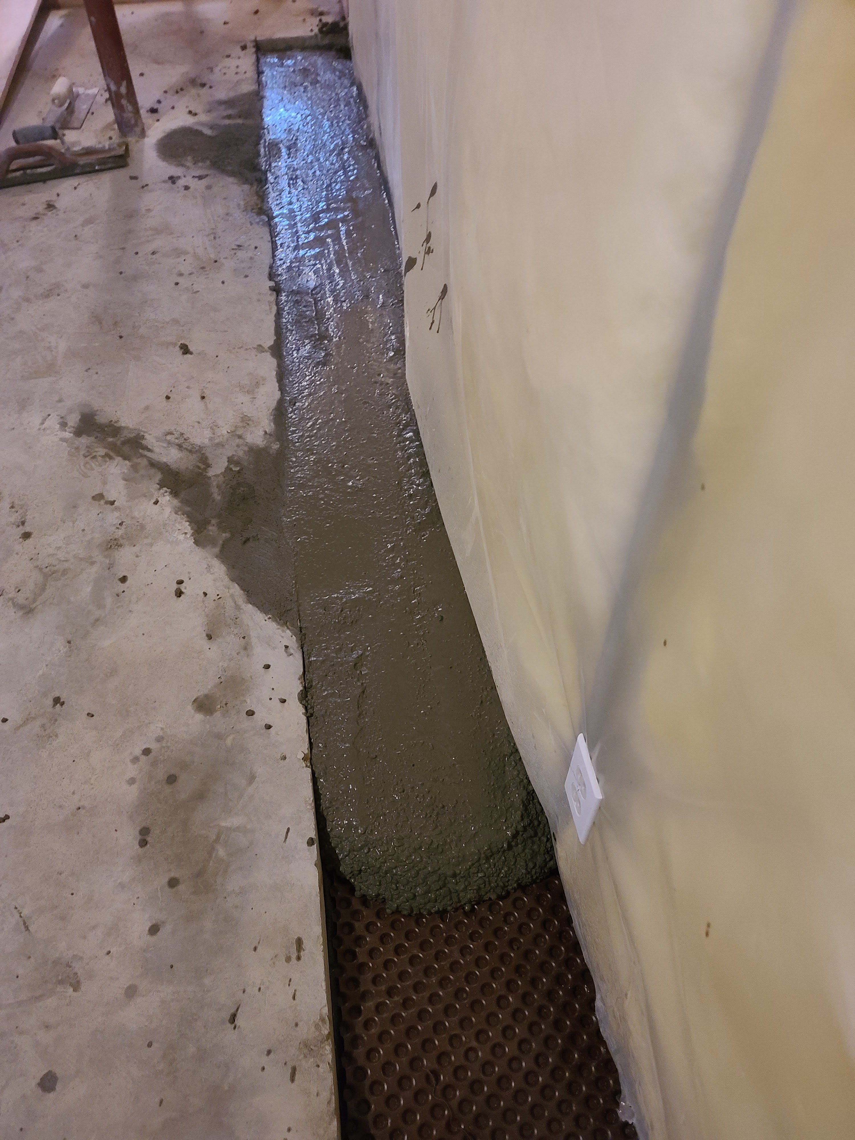 Freshly Poured Concrete Patch Along The Edge Of A Basement Floor Next To A Wall With A Drainage Mat Visible Underneath